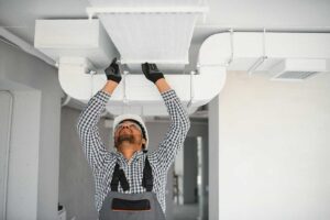 Technician inspecting a residential furnace beside ductwork with tools and a flashlight.